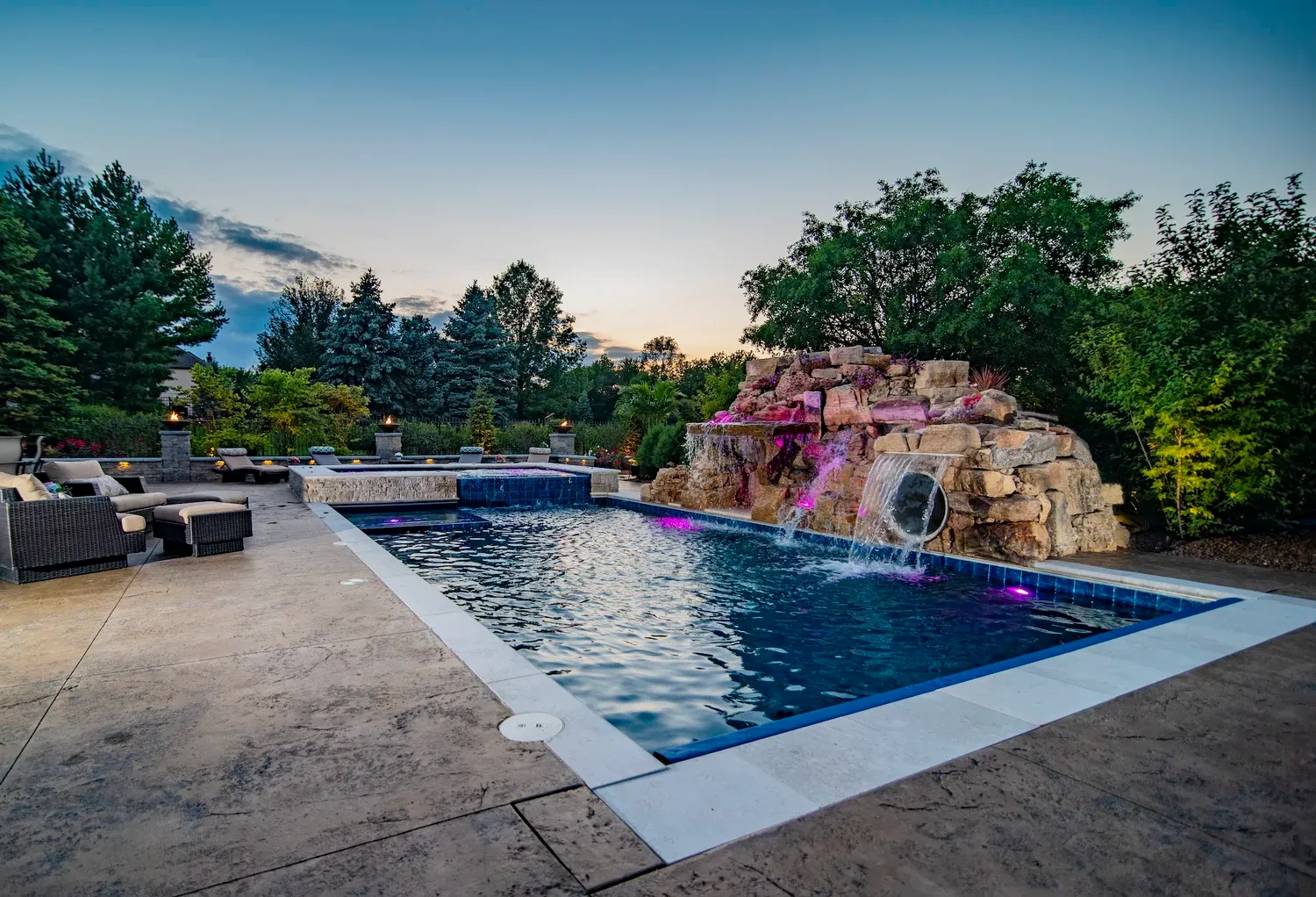 Illuminated custom pool with artisan rock waterfall, spa, and outdoor living patio at twilight in Spring Hill, FL