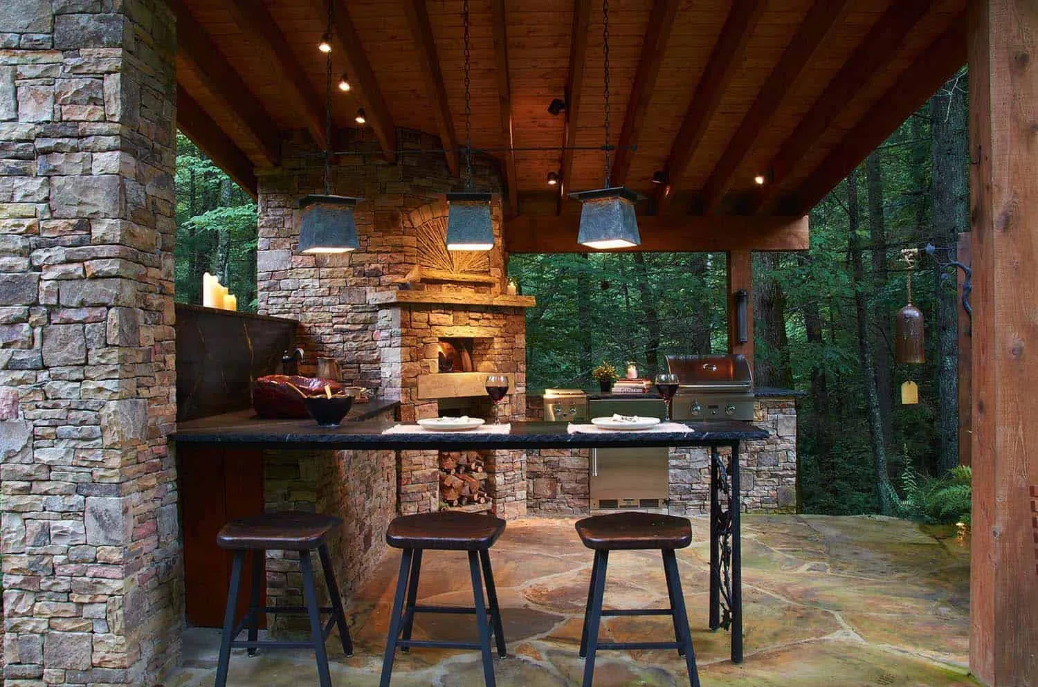Luxurious outdoor kitchen with stone fireplace, bar, grill, and forest views