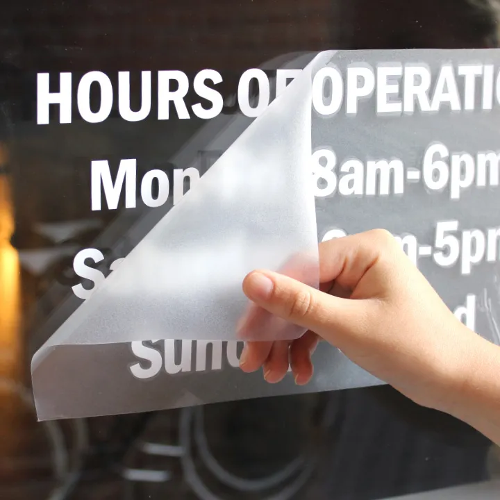 Hand peeling a vinyl decal from a glass surface displaying operating hours.