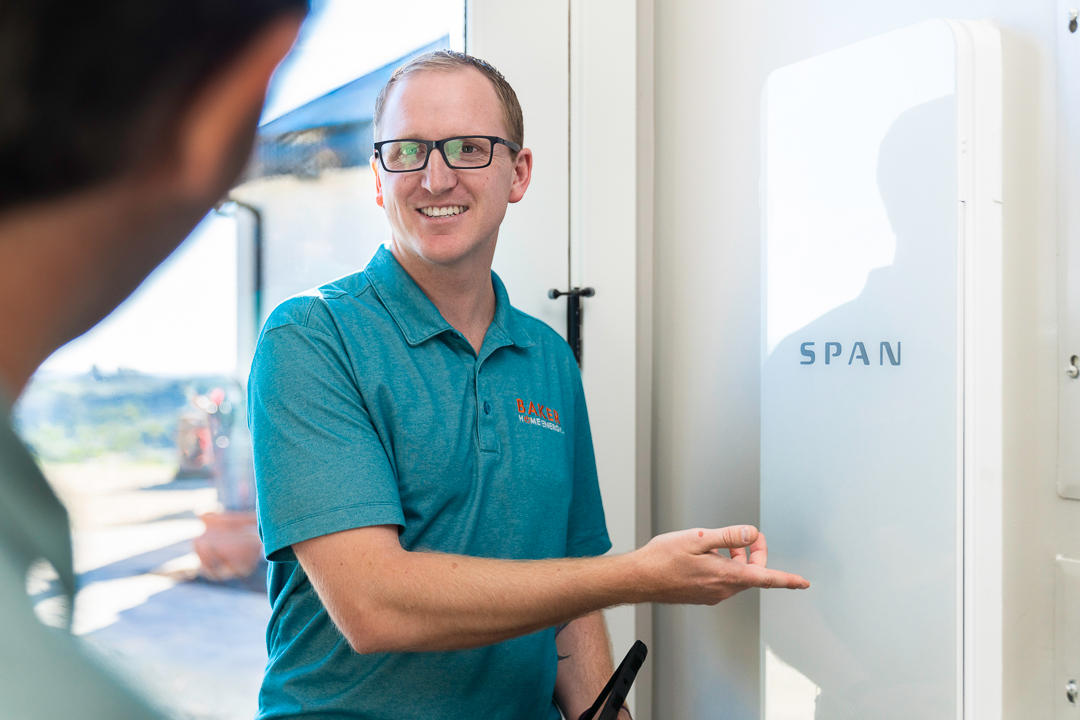 A smiling man in a polo shirt points to a white SPAN energy panel, explaining its features to another person